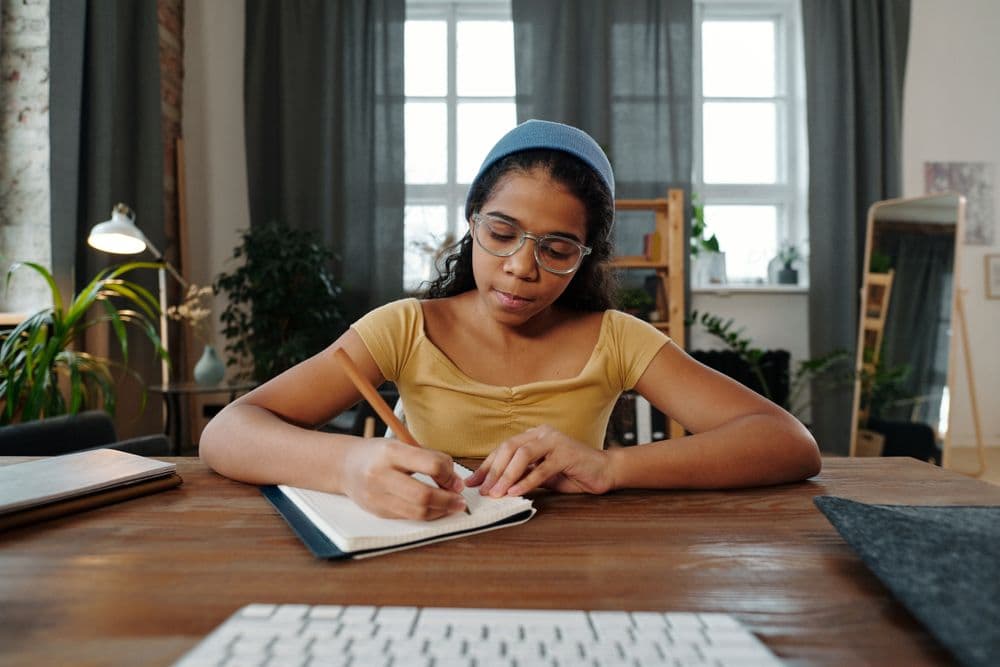 A teenage student writes notes in a notebook at a desk at home